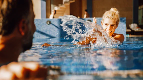 A woman smilingly splashes water in the direction of a man. Both are swimming in the pool, with other people on loungers in the background. 
