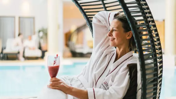 A woman in a white robe sitting in a lounge chair by a pool, holding a drink in her hand. 