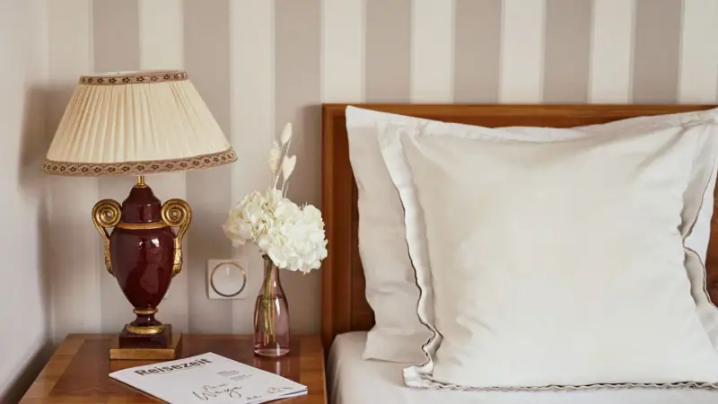 A lamp on a nightstand next to a bed with cushions and linens.
