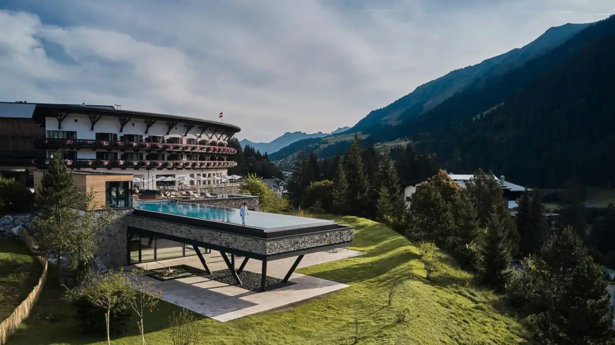 Außenansicht Ifen Hotel Kleinwalsertal mit Infinity Pool Ein Gebäude mit einem Infinity Pool auf Stelzen in der Mitte eingebettet in eine grüne Berglandschaft..