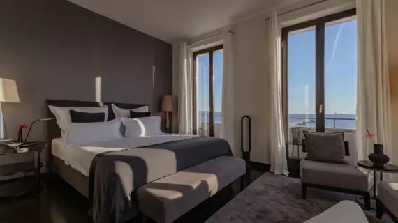 An elegant bedroom with a view of the sea in the sunshine. In the center of the room is a large bed with grey bed linen and white and dark grey pillows. There are bedside tables with table lamps on either side of the bed and a comfortable seat next to the bed.