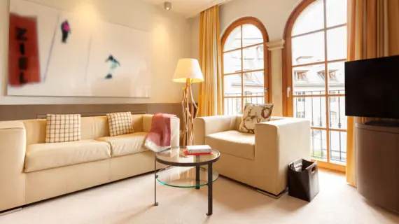 A light-flooded living area with a sofa and an armchair in light-colored leather. In front of the sofa and armchair is a small round glass table with two books on it. Two floor-to-ceiling windows and a television can be seen in the background.