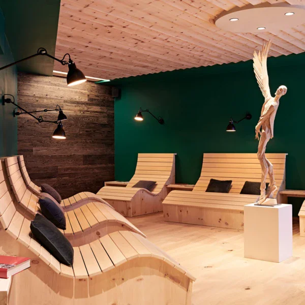 A relaxation room with dark green walls and wooden floor. Along the walls are several wooden loungers with black reading lamps attached to the walls and in the centre of the room is a bright figure of a man with wings. 