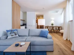 A living area with a gray sofa in front of a wooden coffee table. An open kitchen and a dining table with six chairs can be seen in the background.