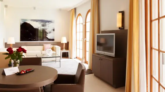 A bright living area with a round wooden table on which stands a vase of red roses and a bowl of fruit. There are also two brown armchairs in the background, a television and a light-colored sofa. 