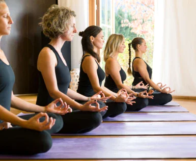 A group of women sitting in a row, wearing yoga pants and practicing yoga indoors.