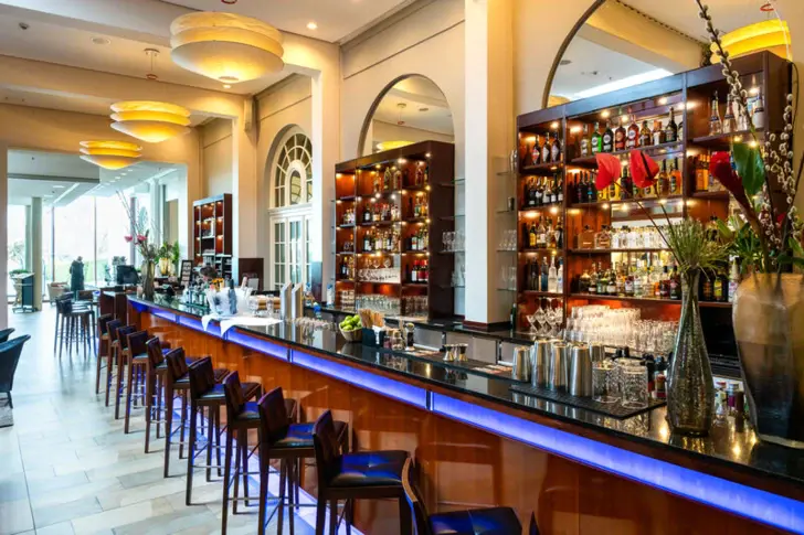 An elegant, well-lit bar area with a blue neon outline under the counter, wooden shelves full of bottles and hanging golden lamps. At the bar counter are high stools with blue seats, and large arched windows can be seen in the background.     User