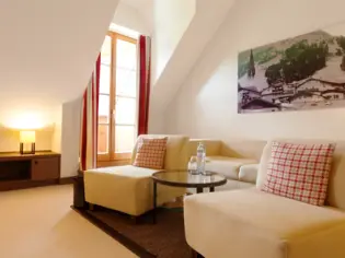 A seating area with a beige sofa and two armchairs with checkered cushions. There is also a small round glass table between the armchairs, on which two glasses and a bottle of water are placed.