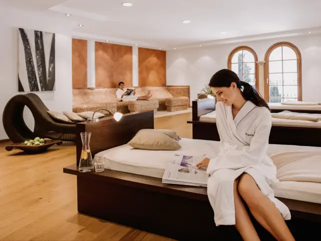 A woman is sitting in a relaxation room with multiple beds, each with their individual reading light and pillows. A man is sitting on a stone lounge in the background. 