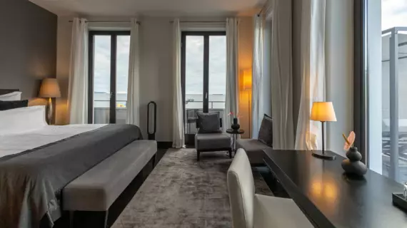 A spacious and stylishly furnished hotel room in daylight. In the foreground is a glossy black desk with a modern chair and decorative objects. Behind it is a large bed with dark gray bed linen and white bed cushions, flanked by an elegant seating area and floor-to-ceiling windows with white curtains and a view of the open sea. 