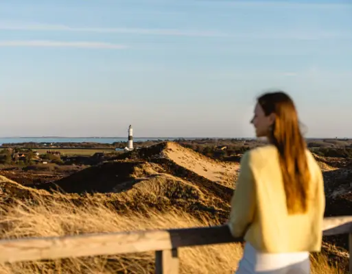 A woman standing on a fence overlooking a lighthouse under a partly cloudy sky.